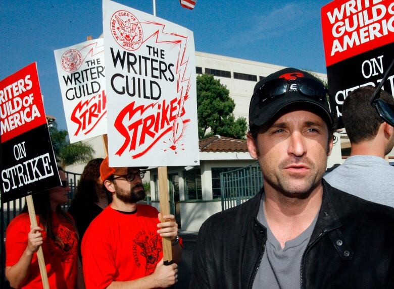 A man wearing a dark baseball hat is surrounded by people holding strike signs.