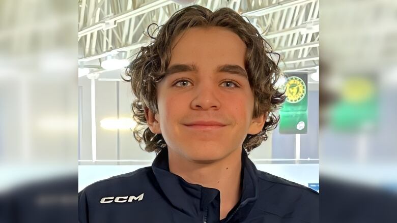 A teen with curly hair looks at the camera while wearing a hockey jacket.