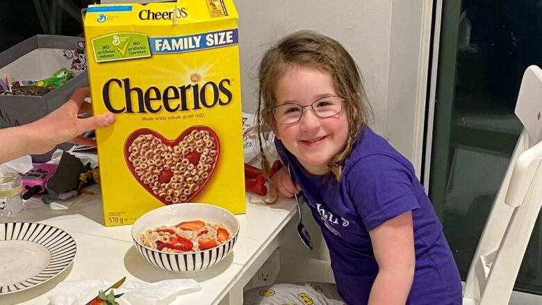 A little girl smiles at the camera. A bowl of cereal is on the table beside her.