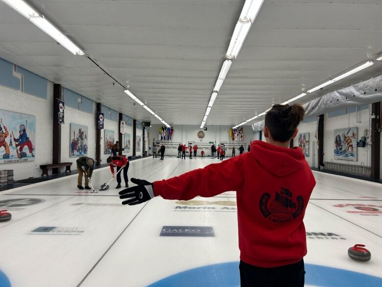 A boy stands on the ice as curlers play a game.