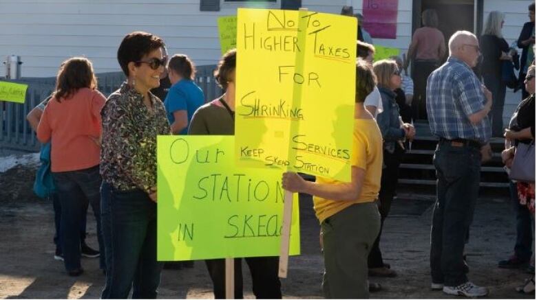 A dozen people stand outside a building in the sunshine holding signs that read 'No to Higher Taxes for Shrinking Services: Keep Skead station' and 'Our Station in Skead' 