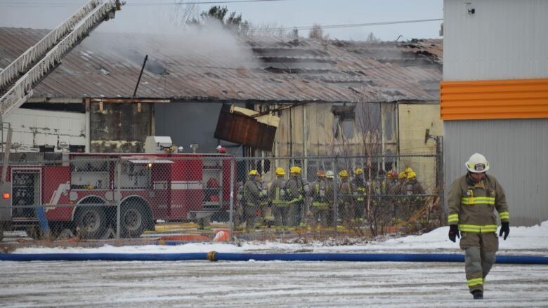 A firefighter walks away from a building with smoke rising from a hole in the roof, with a dozen other firefighters and a fire truck close by