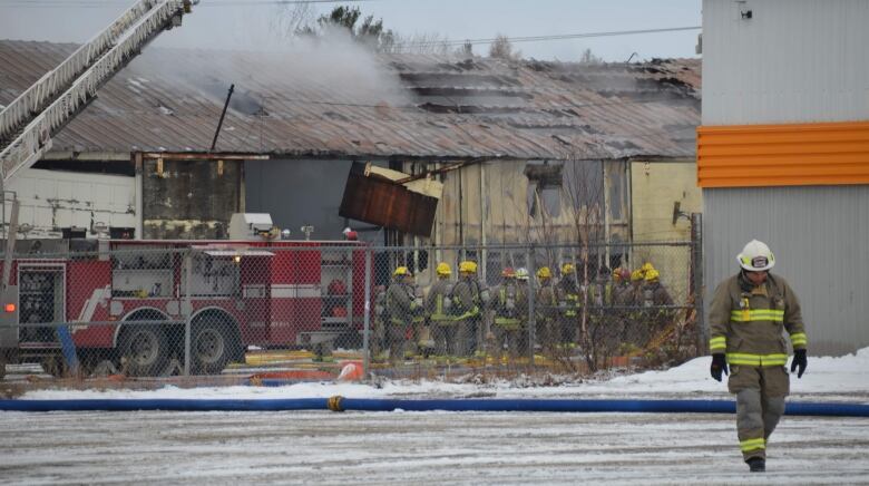 A firefighter walks away from a building with smoke rising from a hole in the roof, with a dozen other firefighters and a fire truck close by