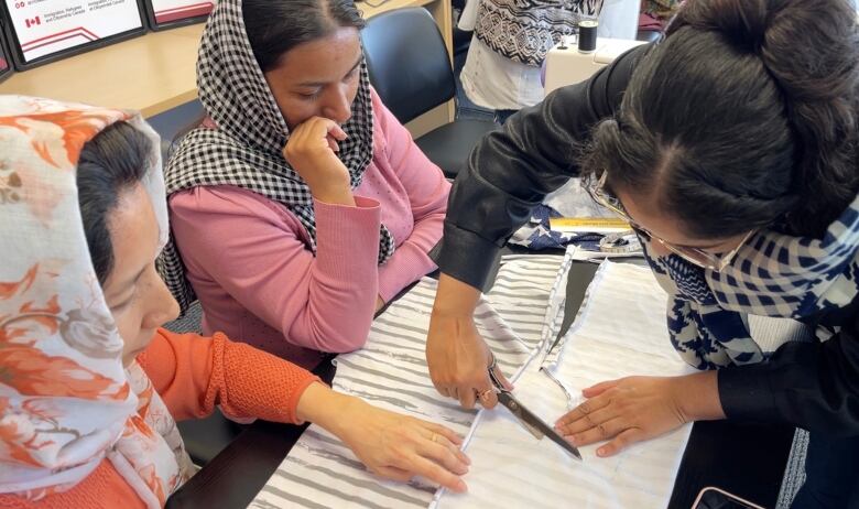 A photo shows three women working with a piece of fabric, one cutting the fabric with scissors.