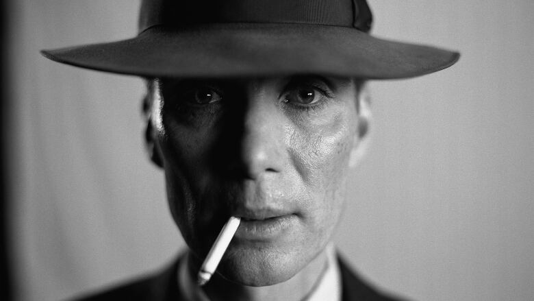 A black and white close up photo of a man looking directly into the camera while wearing a wide brimmed hat and smoking a cigarette. 