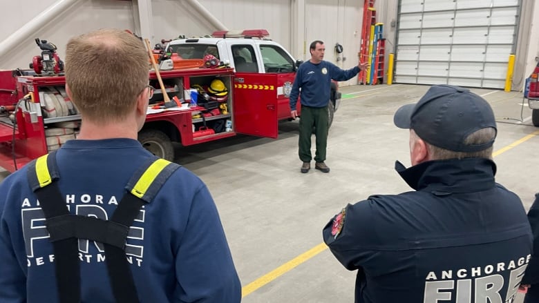 A firefighter stands talking inside a large garage while 2 others look on.