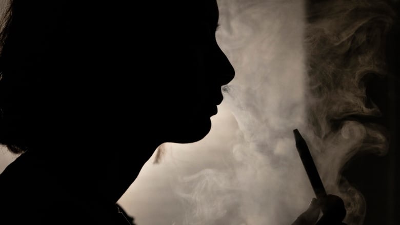 A young person's profile is shown in silhouette as they hold a vape pen in one hand and blow vapour out their nose.