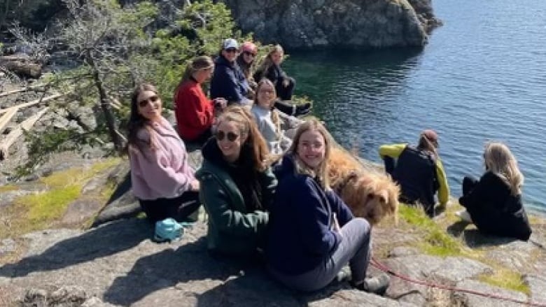 A group of women sit on a rock above the water and mountains. 