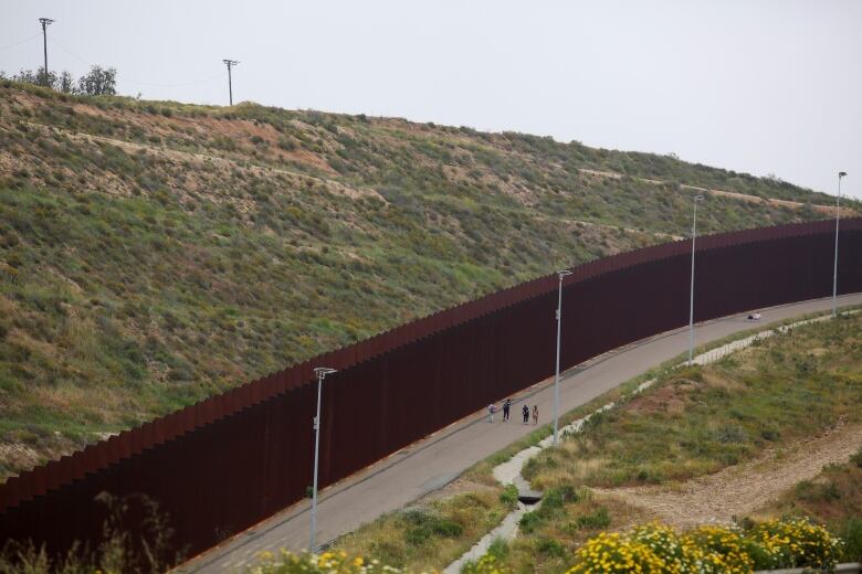 People walk on a trail next to a high, wide fence with a stretch of land on the other side. 