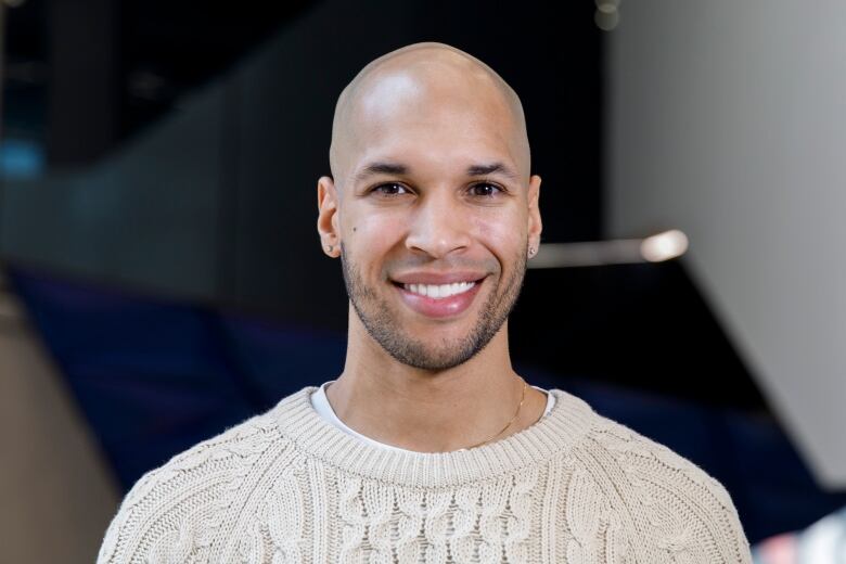 A Black man wearing a cream coloured sweater, thin gold necklace and diamond stud earrings smiles at the camera.   