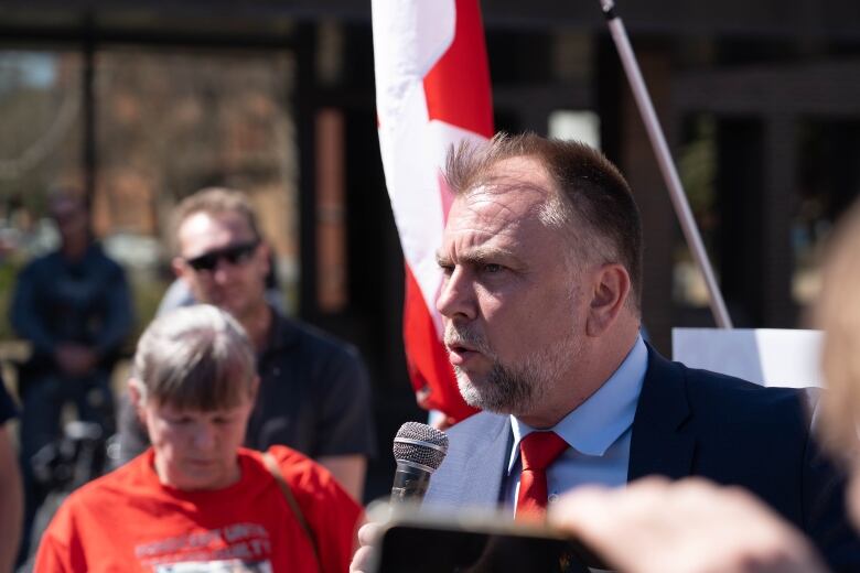 A bearded preacher speaks into a microphone to his supporters. A Canadian flag is behind his head.