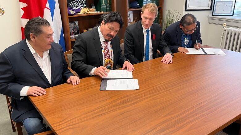 Four men sit at a table in Ottawa, all wearing suits. Two of them sign papers.