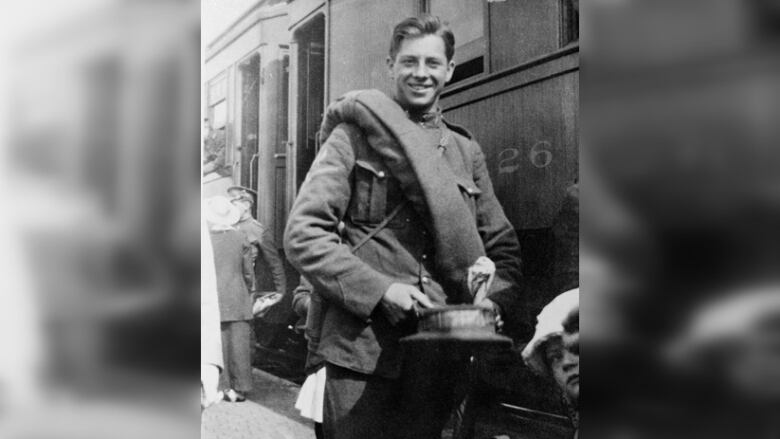 A black and white photo of a smiling soldier in uniform carrying his hat, standing next to a train.