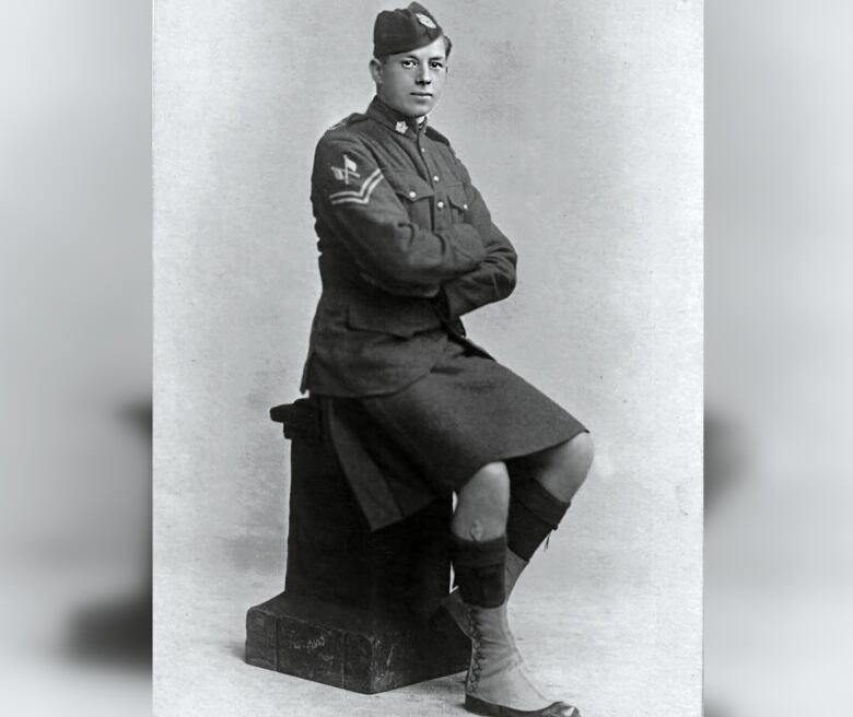 A black and white photo of a soldier in uniform. He's seen sitting on a block with his arms crossed.