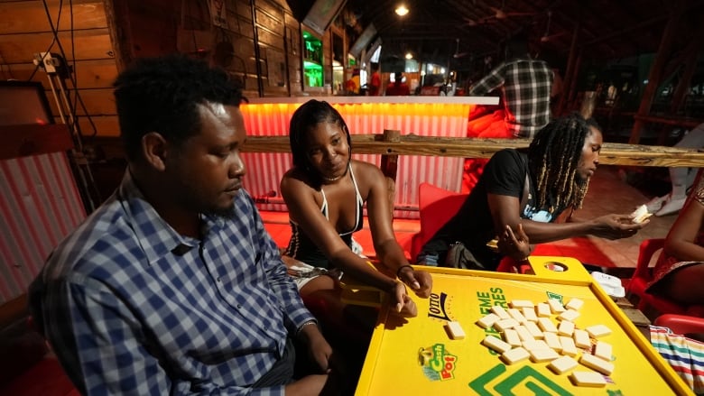 Three people sit around a yellow table, decorated with green logos, playing dominoes in a bar at night. In the background, the main bar is lit up with lights.