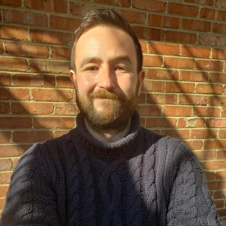 A smiling man wearing a blue sweater stands in front of a brick wall.