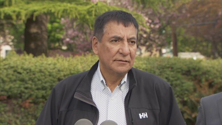 Chief Harley Chingee of the McLeod Lake Indian Band stands in front of greenery in Victoria. He is wearing a black jacket and is speaking in front of several microphones. 