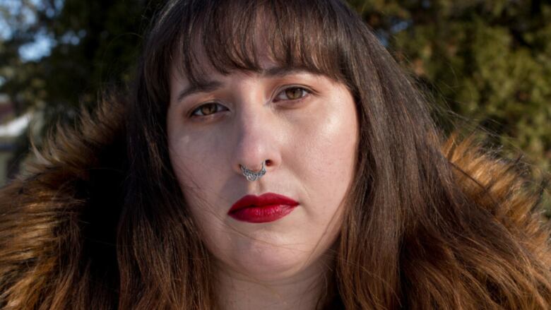 An Indigenous woman with dark brown hair, red lipstick and trees behind her looking at the camera