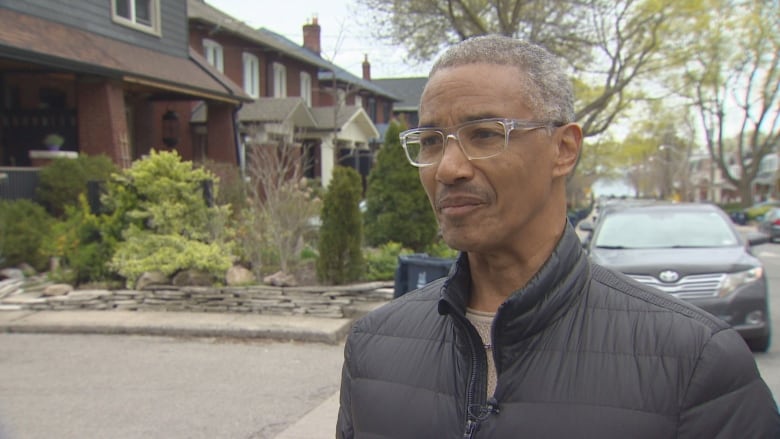 A man with glasses and a black jacket stands on a residential street.