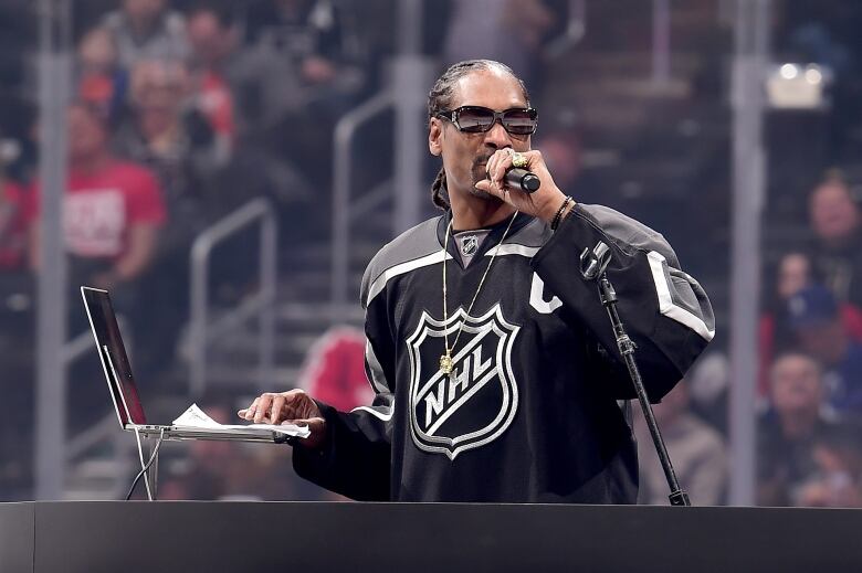 Snoop Dog in an NHL jersey, holding a mic while typing on a laptop with one hand.