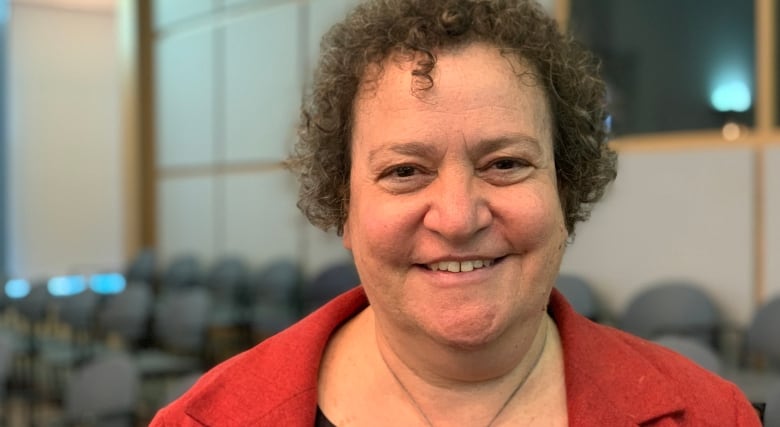 A woman in a red blazer stands in front of some seats and smiles.