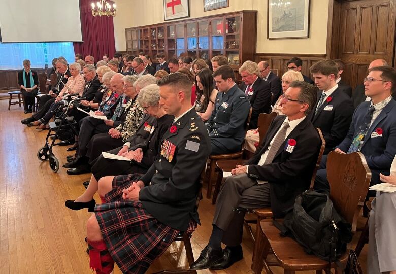 A group of people wearing poppies on their lapels are seated on wooden chairs looking off to the side.