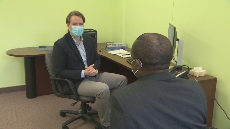 A man wearing a face mask is sitting at a desk talking to another man in an office with light green walls.