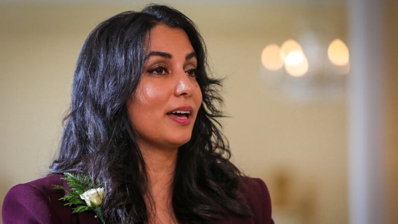 A South Asian woman wearing a mauve jacket and a white flower on her lapel speaks.