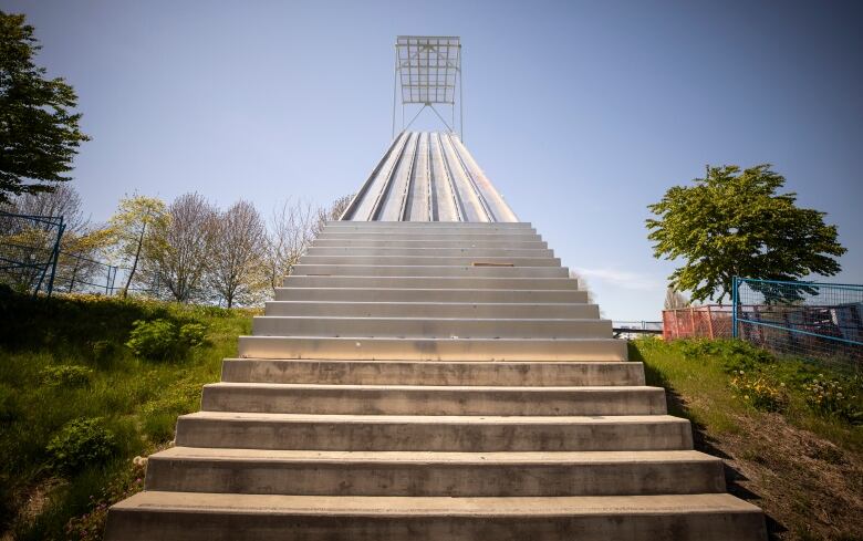 An art piece depicting a series of rising stairs is pictured behind blue fencing.