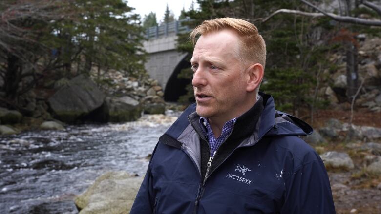 A man in a blue windbreaker stands in front of a river with a bridge in the background.