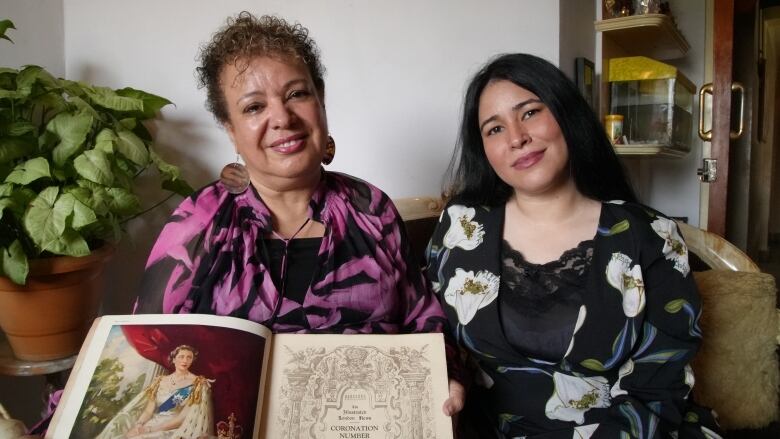 Two women hold a book open to a page showing a coronation illustration of Queen Elizabeth.