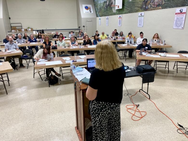 A woman speaks at a podium in front of a room full of people sitting at long tables.