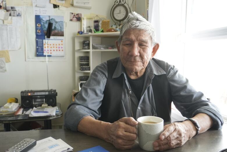 Man with white hair sits next to window holding coffee mug. 