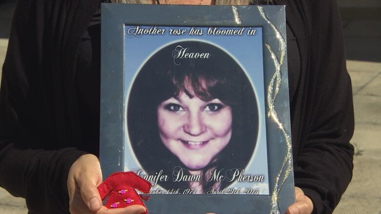 A closeup shows a woman holding a photo of a younger woman in one hand, and a small red beaded pin in the shape of a dress in the other.