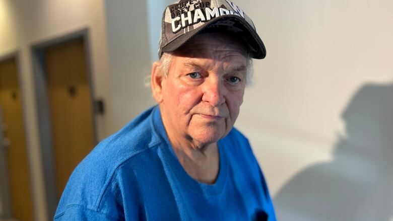 A older man is standing in a hallway wearing a blue shirt and baseball hat. 