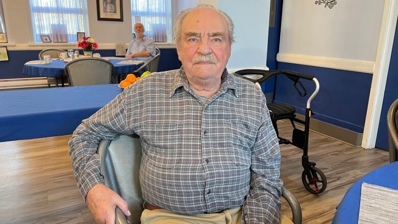 An older man is sitting in a chair in a large room. Tables and a walker can be seen behind him.