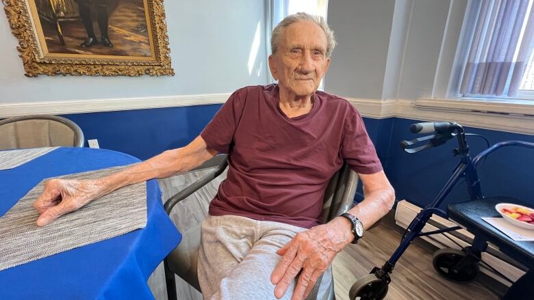 An older man is sitting in a chair at a table. A walker can be seen behind him.