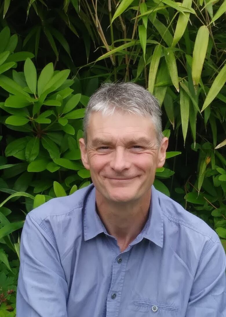 Man in blue shirt smiles in front of greenery