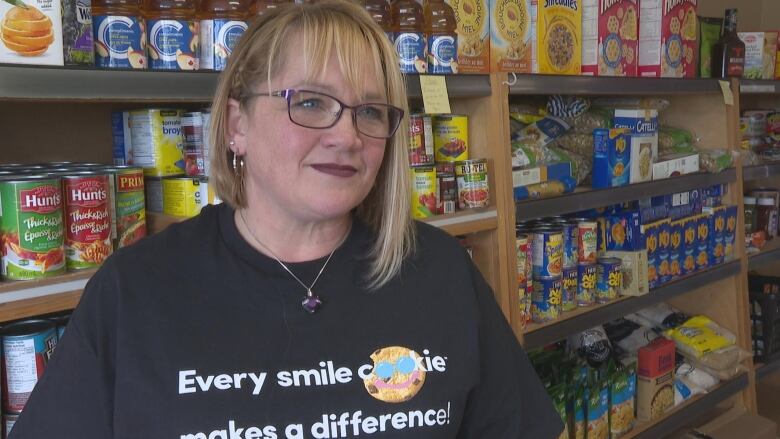 A blonde woman stands in front of shelves of dry and canned food