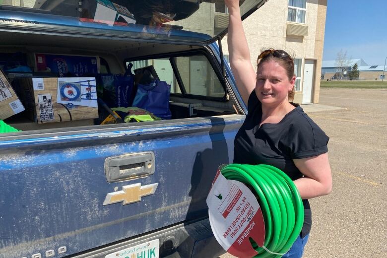 A woman stands holding a garden hose spool in one arm and propping the back winder of a station wagon with her other hand. Inside the vehicle are personal belongings.