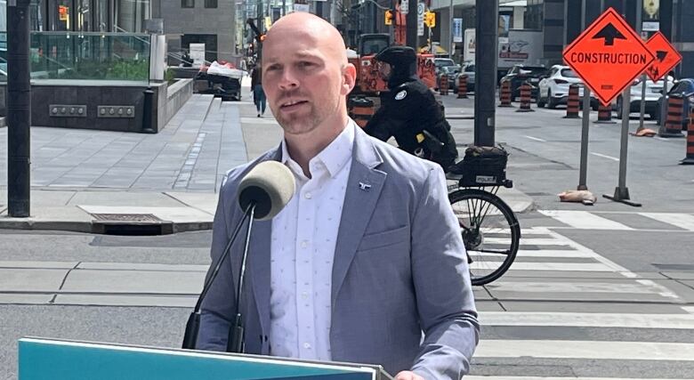 Man in a grey jacket stands behind a podium as vehicles travel through a construction zone behind him.