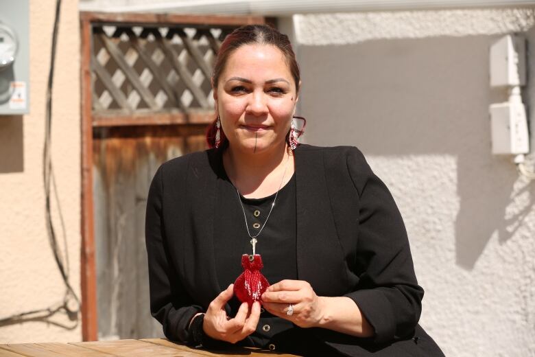 A woman with traditional Inuit face tattoos wearing a black top holds a red beaded dress medallion in front of a wooden door and a white stucco wall.