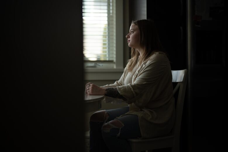 Through a doorway, a woman in a white sweater is seen sitting down at a table and looking out to the left. 