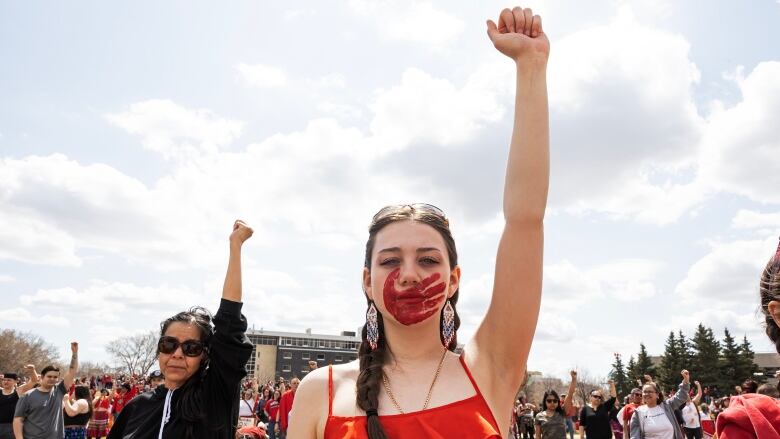 A person in a red tank top with their hair in a long braid and a red hand print over their mouth raises their fist. People standing in the background are doing the same.