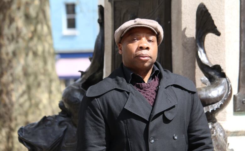 A man wearing a dark-coloured coat and a light-coloured cap stands outside a building.