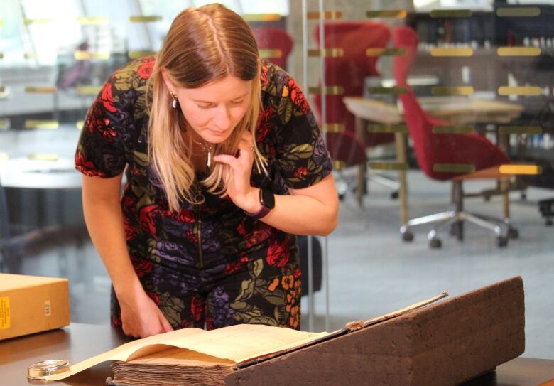 A woman with long blond-brown hair, wearing a floral dress, looks over a thick book.