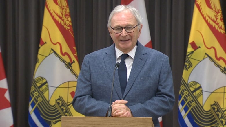 A man with grey hair and glasses, wearing a pale blue blazer, white striped shirt and navy tie, stands at a podium in front of a microphone, with New Brunswick and Canadian flags behind him.