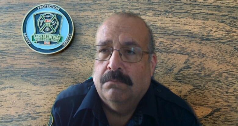 A man with a thick mustache, wearing glasses and a uniform, sits in front of a wood-patterned backdrop with the Saskatchewan Public Safety Agency logo over one shoulder