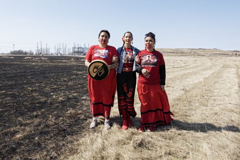 Three women in red skirts stand in a grassy flatland area. One on the left is carrying a hand drum.