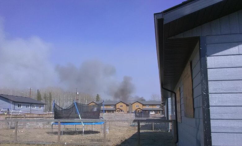 A trampoline stands unused in a backyard with homes around it. Smoke billows in the distance.